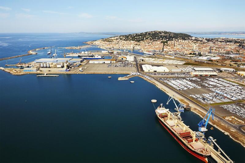 Vue aérienne d’un port industriel en Méditerranée avec navire cargo, grues portuaires, quais logistiques et ville côtière en arrière-plan.