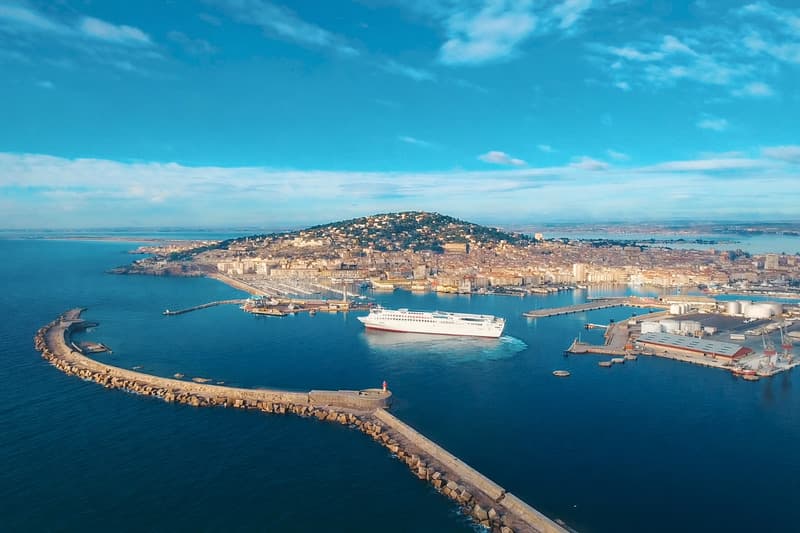 Vue aérienne d’un port méditerranéen avec ferry blanc en manœuvre, digue en pierre, bassins portuaires et ville côtière en arrière-plan.